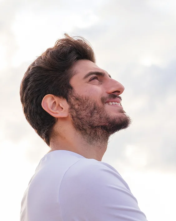 Side profile of a young man with a beard and thick hair, smiling while looking upward, standing outdoors with a cloudy sky behind him - Hair Restoration in New York, NY