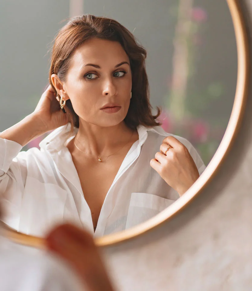 Woman looking at herself in a round mirror while adjusting her hair in a white blouse - Hair Loss in Women in New York, NY